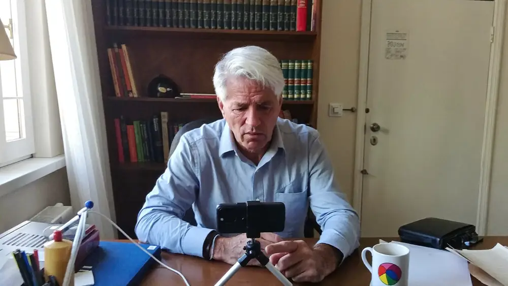 Professor gravando a si mesmo com um celular sobre tripé, em escritório doméstico, sob luz de janela.