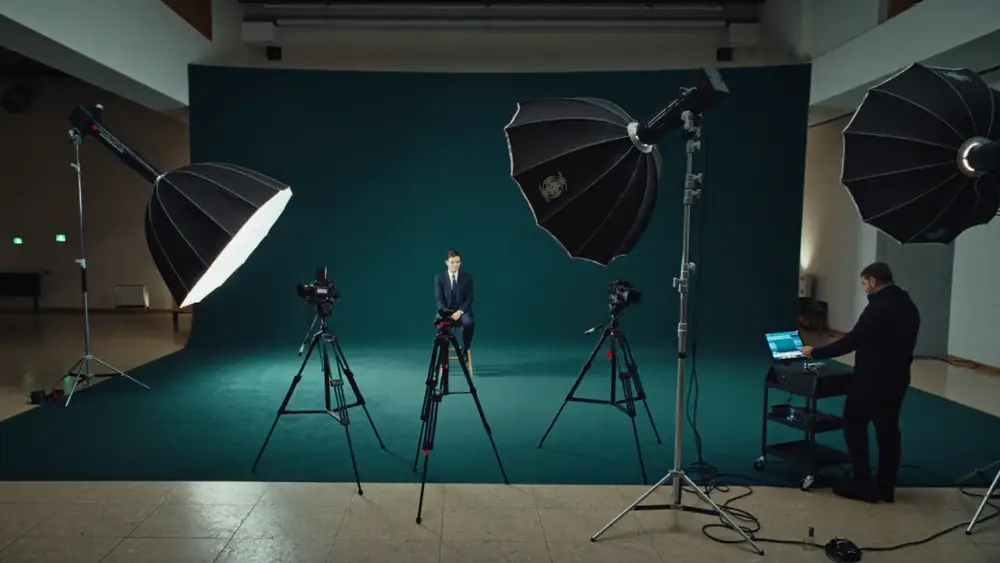 Set de gravação montado dentro de um auditório: duas câmeras em tripé, teleprompter, três softboxes, fundo infinito e um operador conferindo o monitor.